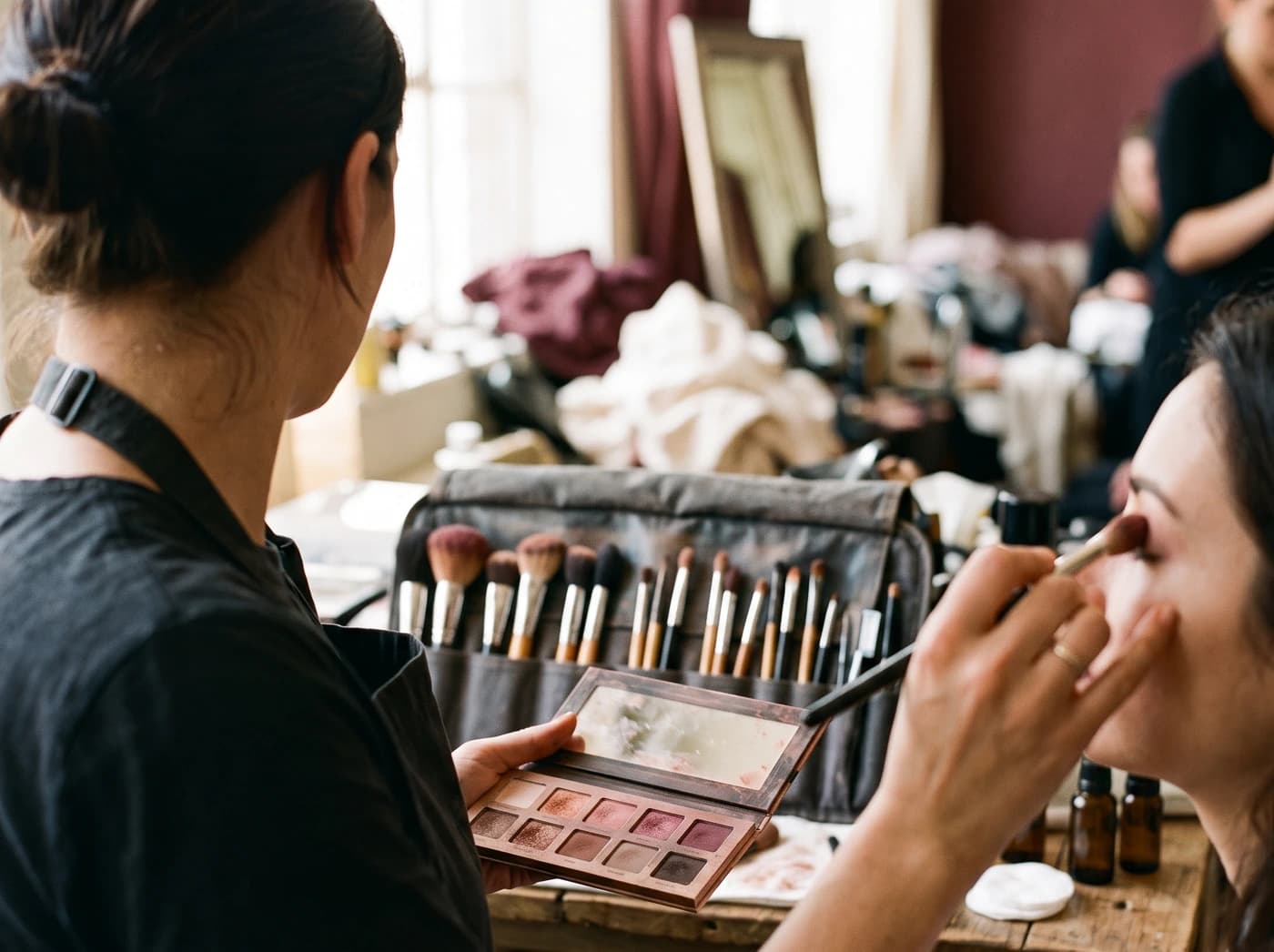 A makeup artist at work, holding an open palette of warm neutral and berry tones and applying colour with a brush, a roll of brushes on the table beside her.